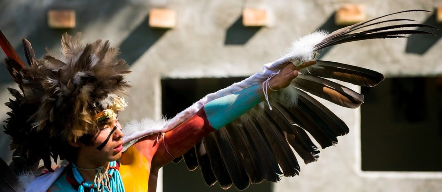 Hopi tribe memeber. His body is painted in bright colors: Blue, teal, and red. He's wearing feathers on his head. His outstretched arm is covered in feathers like the wing of a great bird.